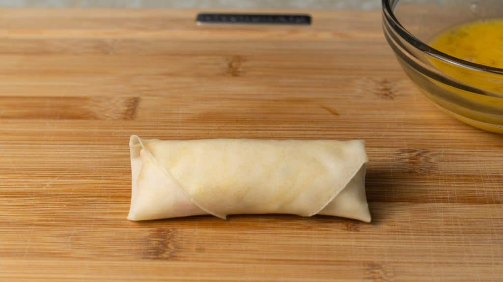 Raw spring roll rolled up on a cutting board beside a bowl of egg wash
