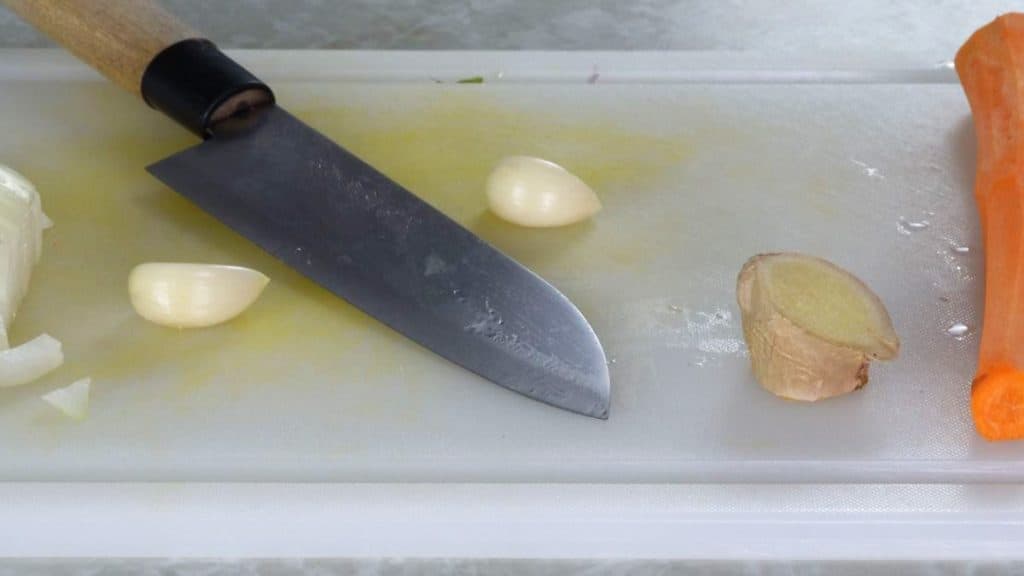 Peeled cloves of garlic on a white cutting board with knife, carrot, and ginger