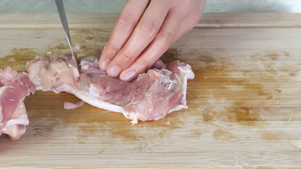 Scoring a raw chicken thigh with a knife on a wooden cutting board