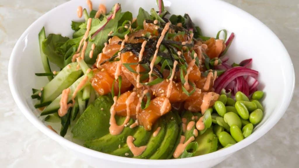 Salmon poke bowl with sliced avocado, edamame, pickled red onion, cucumber, nori, green onions, spring mix, and spicy mayo