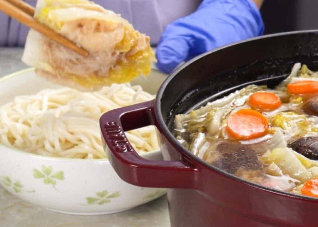 Picking up a piece of napa cabbage and pork belly with chopsticks from a Dutch oven of mille feuille nabe with a bowl of noodles on the side