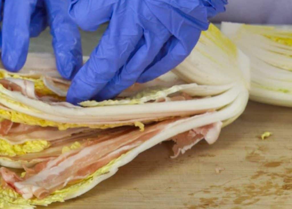 Layering pork belly slices in between napa cabbage leaves on a wooden cutting board