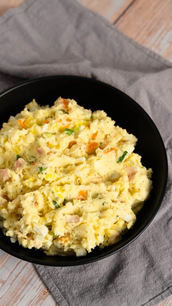 Japanese potato salad with egg, green onions, carrot, potatoes and ham in a black bowl on a gray towel on a wooden surface