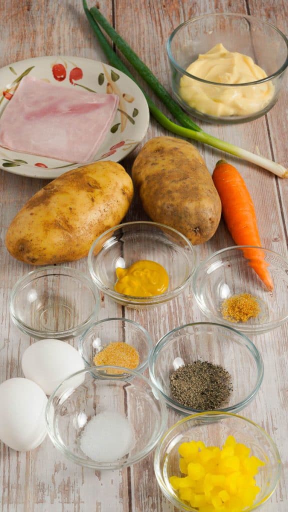 Ingredients for Japanese potato salad, including ham, mayo, green onion, russet potatoes, carrot, mustard, garlic powder, rice vinegar, eggs, black pepper, salt and yellow pickled daikon on a wooden surface