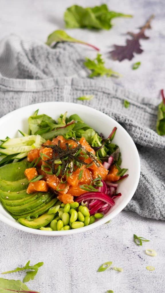 Salmon poke bowl with diced raw salmon, sliced avocado, edamame, pickled red onions, julienned cucumbers and nori seaweed strips in a white bowl 