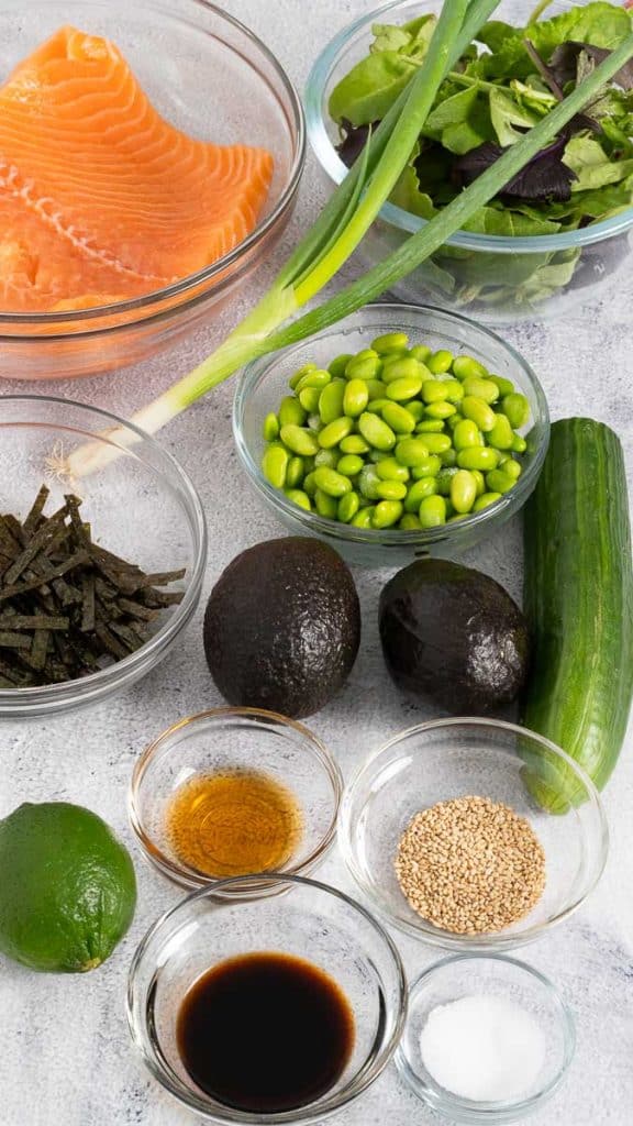 Ingredients for salmon poke bowl including raw salmon, spring mix, green onion, nori, avocadoes, edamame, cucumber, lime, soy sauce, sesame seeds, sesame oil, and salt