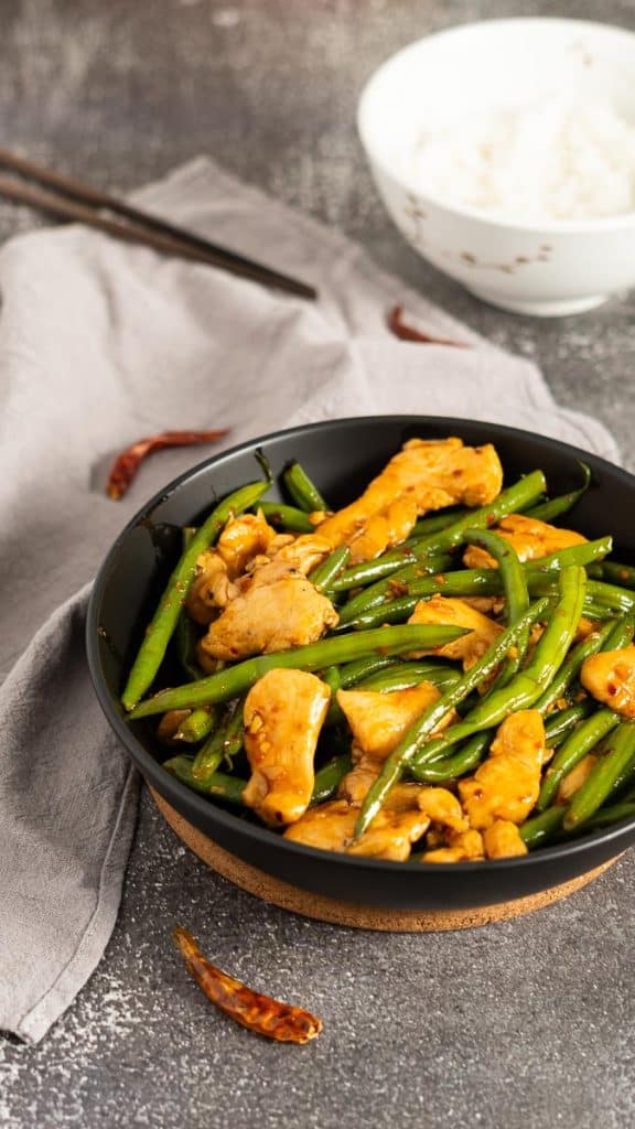 Spicy chicken and green beans stir-fry served in a black bowl on a rustic surface with a bowl of steamed rice