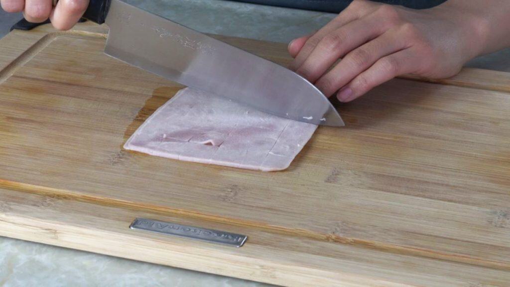 Slicing cooked ham into small squares with a knife on a wooden cutting board