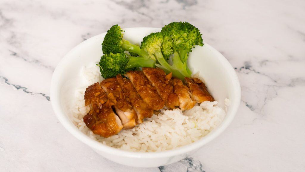 Teriyaki chicken on top of steamed white rice with broccoli on the side on a marble surface
