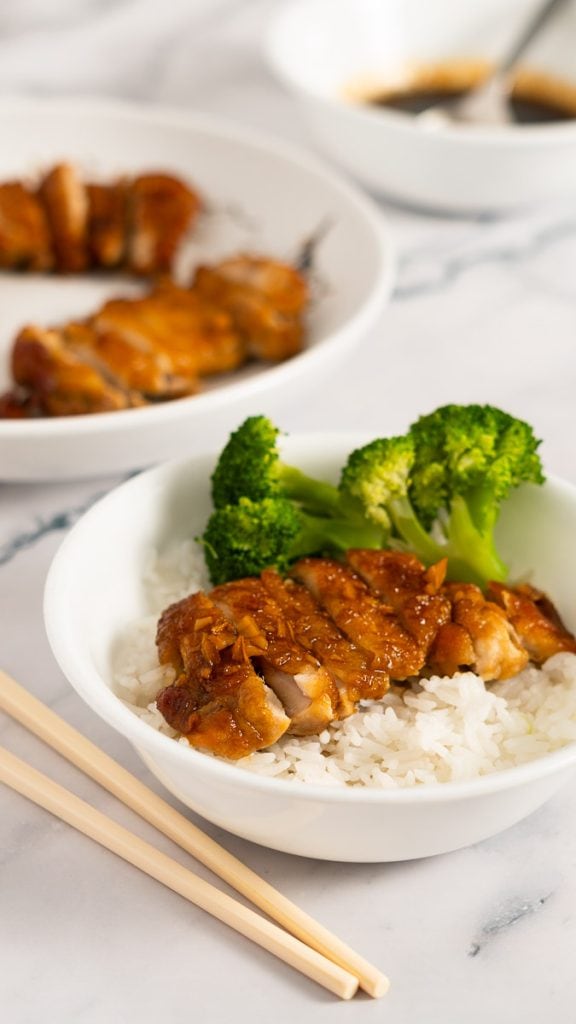 Sliced teriyaki chicken with broccoli on steamed white rice in a  white bowl on a marble surface