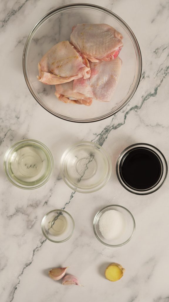 Ingredients for teriyaki chicken, including raw chicken thighs, mirin, sake, soy sauce, rice vinegar, sugar, garlic and ginger