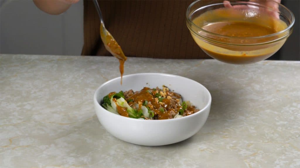 Drizzling spicy sauce with a spoon in a small bowl of noodles with bok choy, chopped peanuts, ground pork and green onions on top