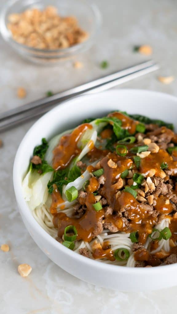 Bowl of wheat noodles with ground pork, bok choy, spicy sauce, chopped peanuts and green onions on top with a small bowl of chopped peanuts