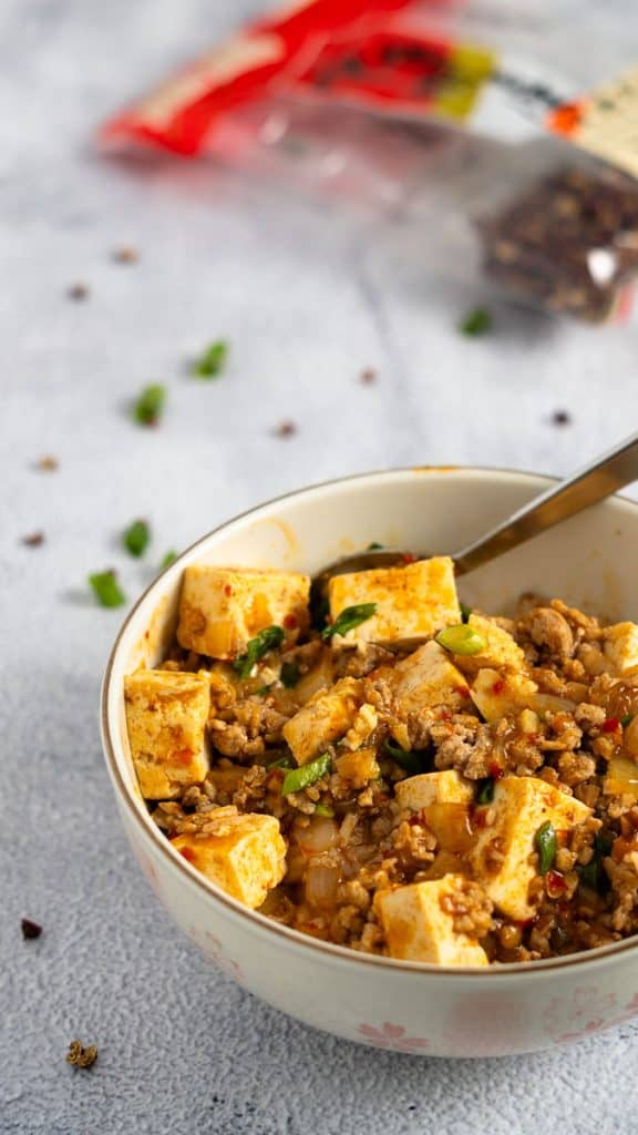 Mapo tofu in a bowl with a bag of Sichuan peppercorns and green onions scattered across the table