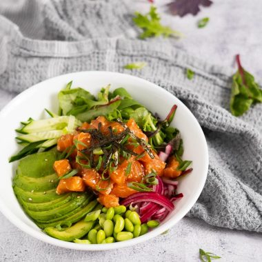 Salmon poke bowl with sliced avocado, cucumber, pickled red onion, spring mix, edamame, nori strips, green onion and sesame seeds in a white bowl on a rustic surface with a gray towel