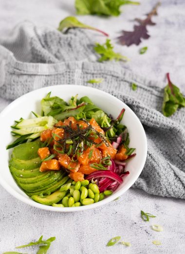 Salmon poke bowl with sliced avocado, cucumber, pickled red onion, spring mix, edamame, nori strips, green onion and sesame seeds in a white bowl on a rustic surface with a gray towel