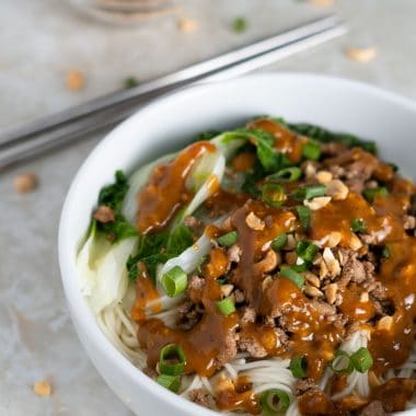 Small bowl of noodles with spicy sauce, baby bok choy, ground pork, chopped peanuts and green onion