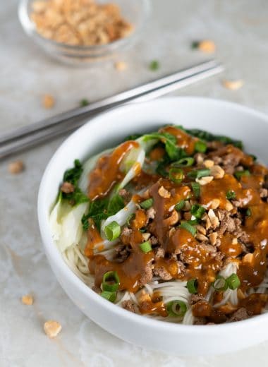 Small bowl of noodles with spicy sauce, baby bok choy, ground pork, chopped peanuts and green onion